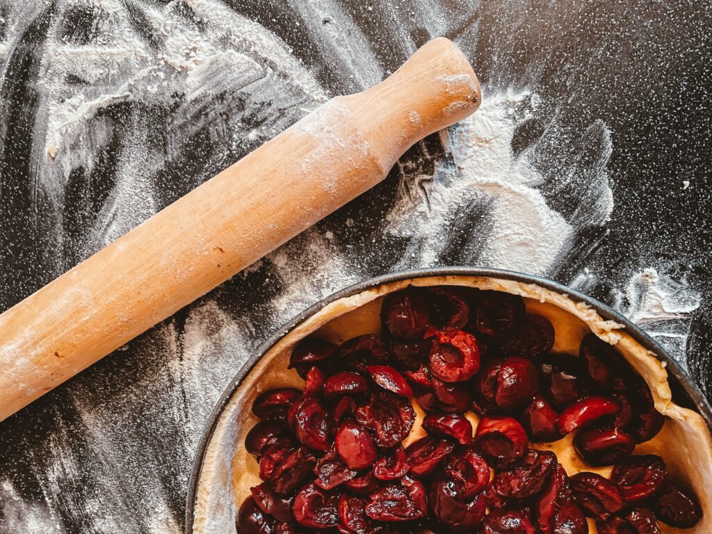 Foto in cui abbiamo un piano di lavoro nero con della farina, un mattarello e la teglia in cui c'è la crostata e il ripieno di ciliegie.